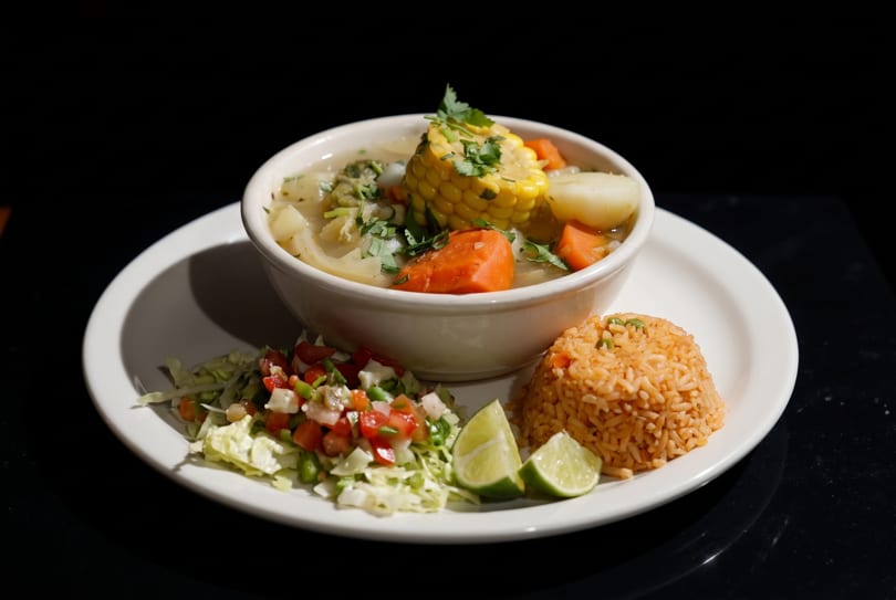 Hearty soup with corn, carrots, and potatoes, served with rice, salad, and lime on a white plate.