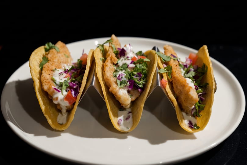 Three crispy fish tacos with white sauce, red cabbage, tomatoes, and cilantro in a metal holder.