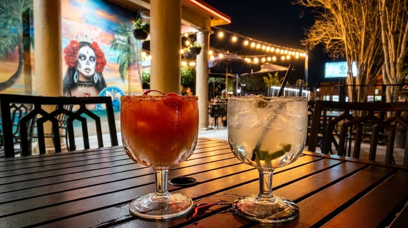 Two cocktails on a wooden table at night, with string lights and a mural in the background.