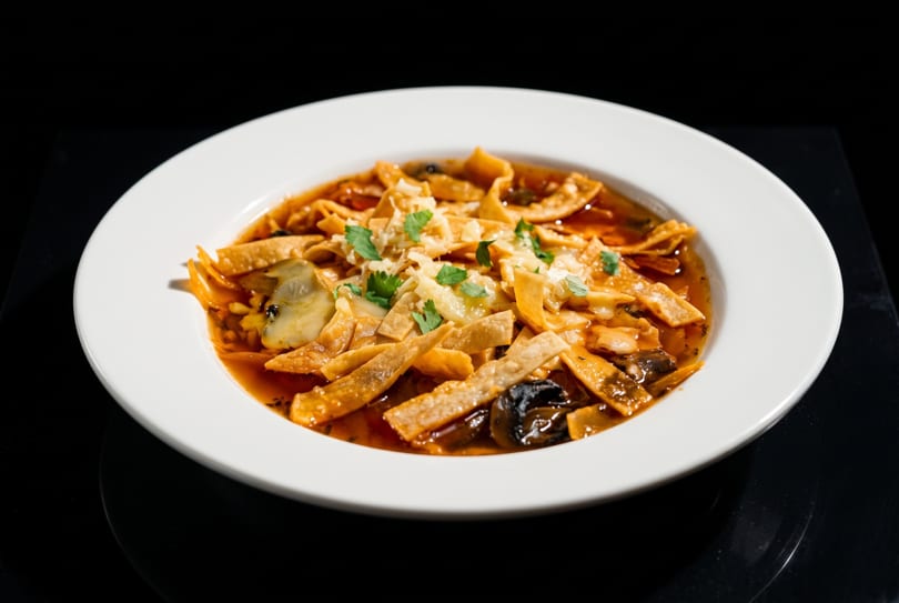 Savory tortilla soup with melted cheese, cilantro, and dark vegetables in a white bowl.