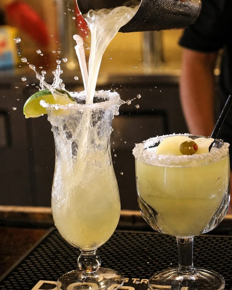 Margarita being poured into a salt-rimmed glass with a lime, next to another margarita.
