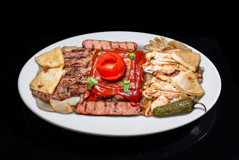 A white platter with grilled steak, chicken, sausage, onions, tortillas, and a tomato-topped sauce.