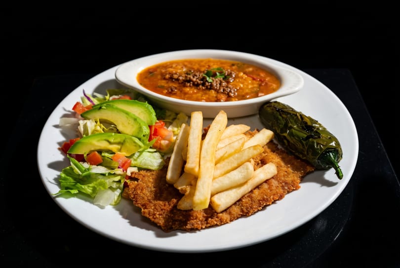 Fried chicken milanesa with french fries, avocado salad, a roasted pepper, and a bowl of soup.